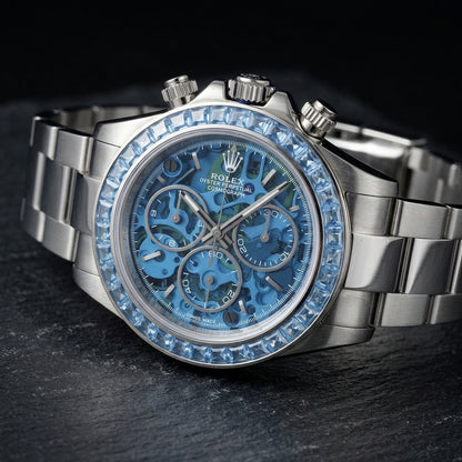 Steel skeleton chronograph at a three-quarter angle on dark slate, the sky-blue stones beside the fluted crown and pushers