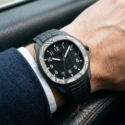 Forged-carbon watch with a black dial on its black rubber strap, worn on the wrist below a dark suit cuff