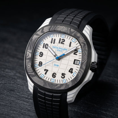 White carbon grid-dial watch at a three-quarter angle on dark slate, grey-black layer lines along the white case flank, a black fluted crown and the black rubber strap