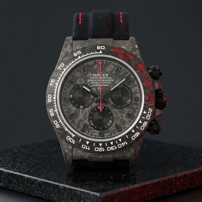 Forged-carbon chronograph on a black woven strap with red stitching, worn under a cuff