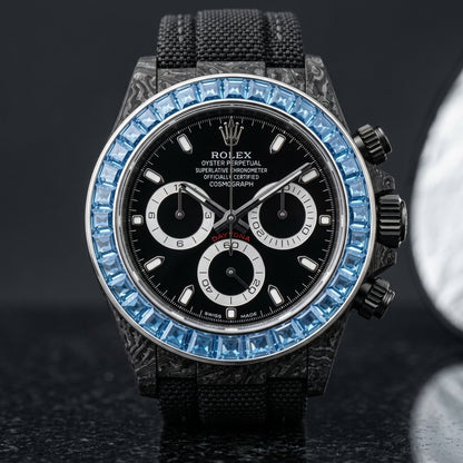 Black forged-carbon chronograph with a sky-blue stone bezel and a black dial, upright on polished black granite