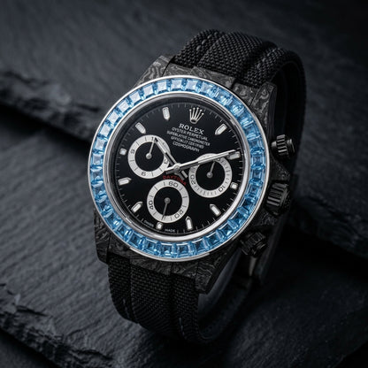 Black forged-carbon chronograph at a three-quarter angle on dark slate, swirling grey grain on its flank and black pushers beside the blue stones