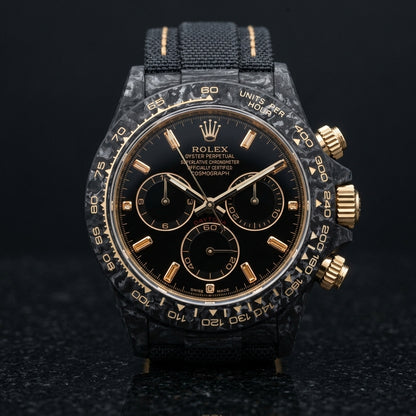 Black forged carbon chronograph with champagne baguette stone markers on a black dial and a pale gold tachymeter scale, upright on polished black granite