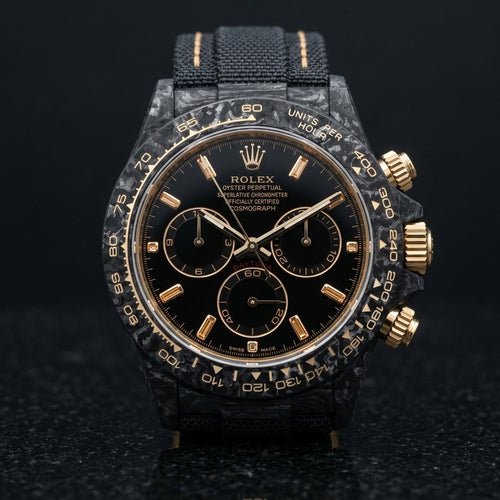 Black forged carbon chronograph with champagne baguette stone markers on a black dial and a pale gold tachymeter scale, upright on polished black granite