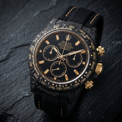 Black forged carbon chronograph at a three-quarter angle on dark slate, its gold crown and pushers against the marbled carbon flank