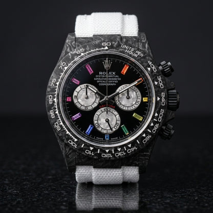 Black carbon chronograph with rainbow stone markers on its white woven strap, worn on the wrist below a dark suit cuff