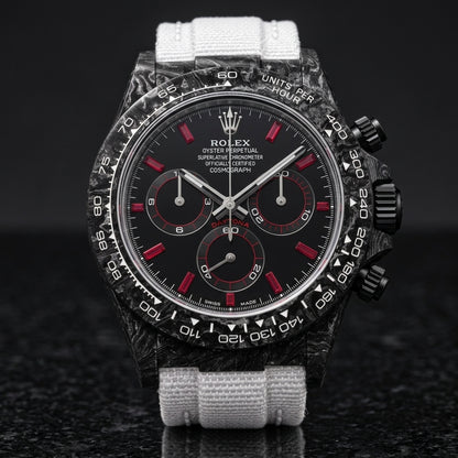 Black carbon chronograph with red baguette markers on its white woven strap, worn on the wrist below a suit cuff