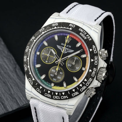 White forged carbon chronograph at a three-quarter angle on dark slate, its rainbow minute track inside the black marbled bezel