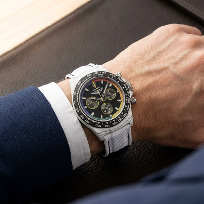White carbon rainbow-ring chronograph on its white woven canvas strap, worn on the wrist below a navy suit cuff
