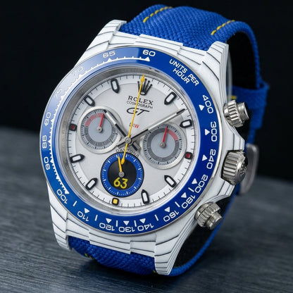 White forged-carbon chronograph at a three-quarter angle on dark slate, the blue bezel beside the steel crown with its yellow ring