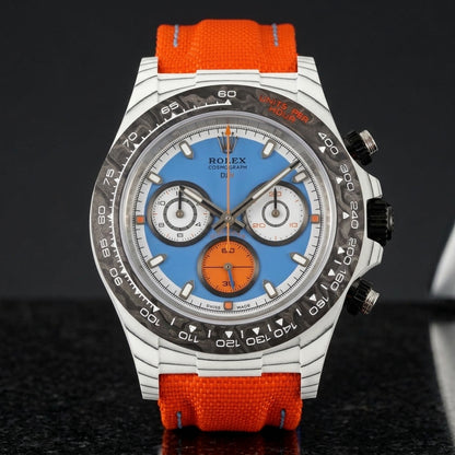 White carbon chronograph with a sky-blue dial, an orange counter and a black forged-carbon bezel, upright on polished black granite