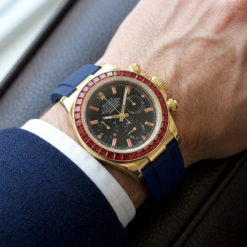 Gold chronograph with a red stone bezel on its navy blue rubber strap, worn on the wrist below a dark suit cuff