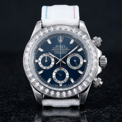 Steel chronograph with a blue dial on its white canvas strap with sky-blue and pink stitching, worn on the wrist below a dark suit cuff