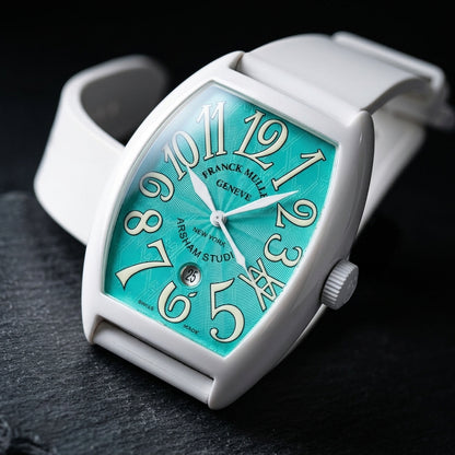 White curved tonneau watch at a three-quarter angle on dark slate, the curve of its white flank and its fluted white crown beside the turquoise dial
