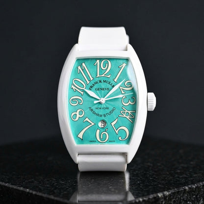 White curved tonneau watch with a turquoise dial on its white rubber strap, worn on the wrist below a dark suit cuff