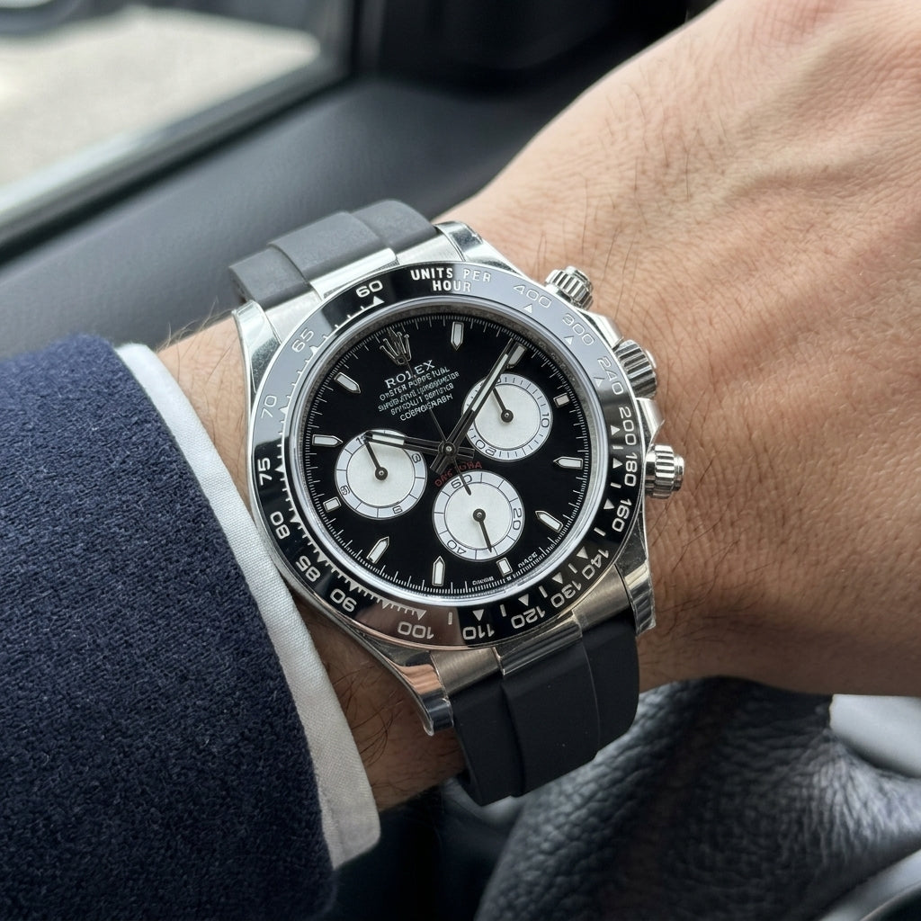 Steel chronograph with a black dial and white counters on its black rubber strap, worn on the wrist below a dark suit cuff