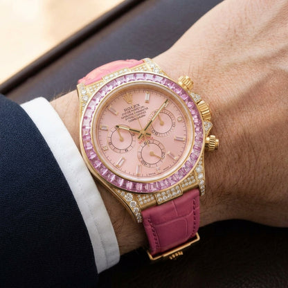 Gold chronograph with a pink dial and a pink stone bezel on a pink alligator strap, worn on the wrist below a navy suit cuff