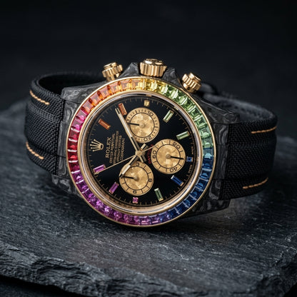Black forged carbon chronograph at a three-quarter angle on dark slate, its gold crown and pushers beside the rainbow bezel