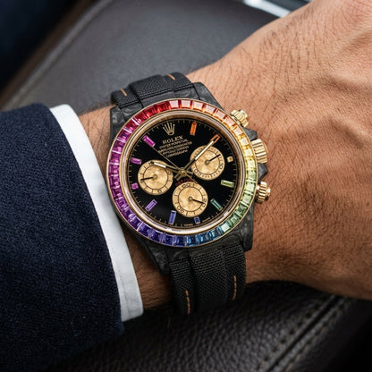 Black carbon chronograph with a rainbow bezel on its black woven strap with tan stitching, worn on the wrist below a dark suit cuff