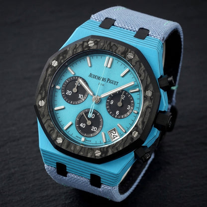 Octagonal chronograph at a three-quarter angle on dark slate, black layer lines through the sky-blue flank beside the black crown and pushers