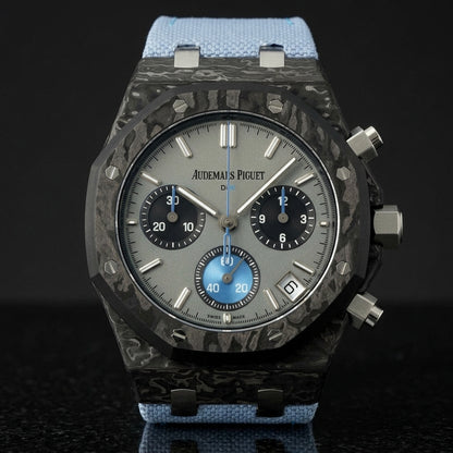 Black forged-carbon octagonal chronograph with a grey dial and a sky-blue counter, upright on polished black granite
