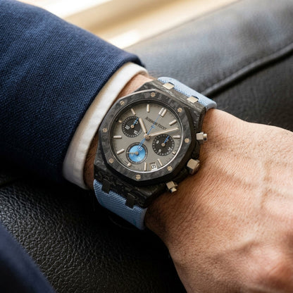Black forged-carbon octagonal chronograph on its light-blue canvas strap, worn on the wrist below a dark suit cuff