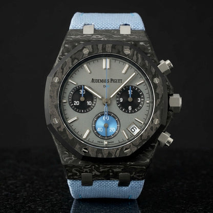 Black forged-carbon octagonal chronograph on its light-blue canvas strap, worn on the wrist below a dark suit cuff