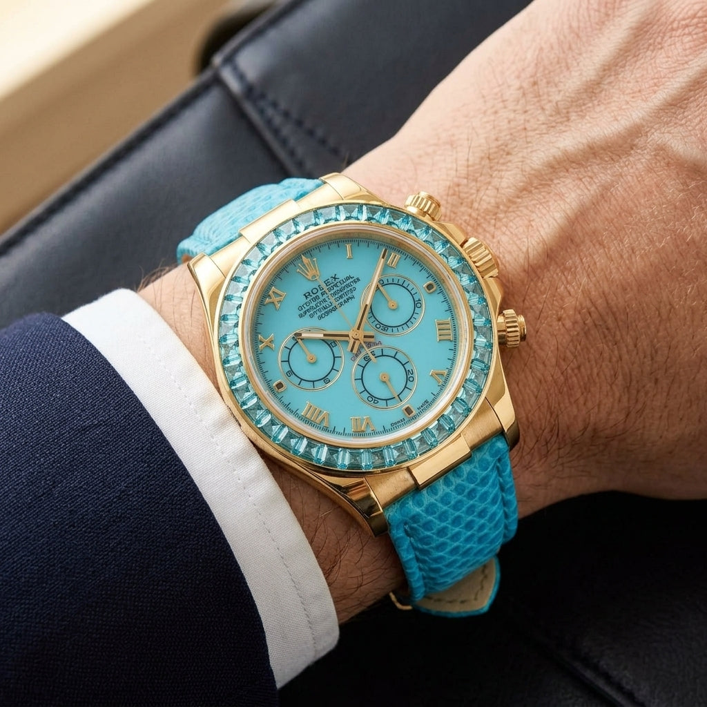 Gold-tone chronograph with a turquoise dial and an aqua-blue stone bezel on a turquoise lizard-grain strap, worn on the wrist below a navy suit cuff