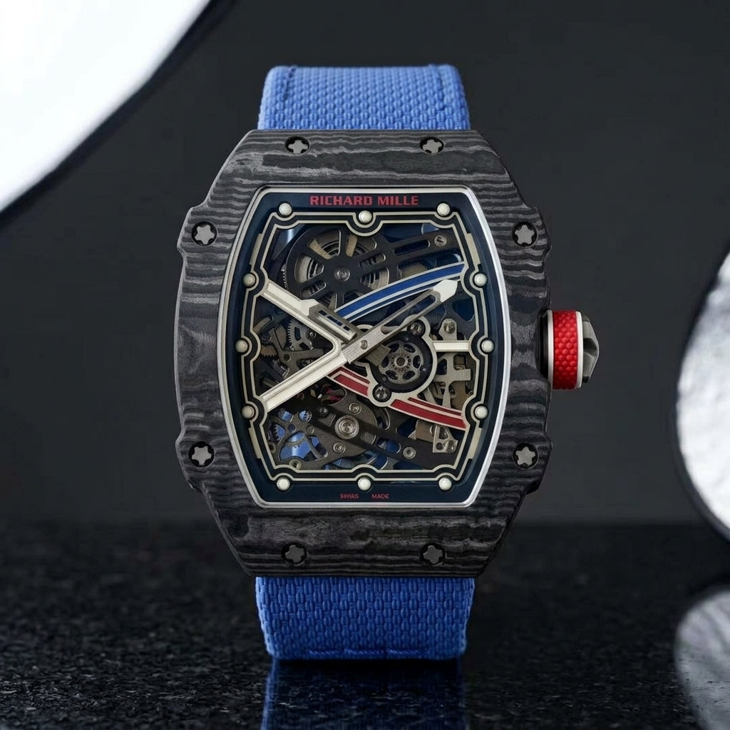 Black carbon tonneau watch with a red crown on a blue textile strap, worn on the wrist below a navy suit cuff
