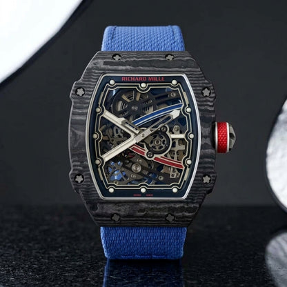 Black carbon tonneau watch with a red crown on a blue textile strap, worn on the wrist below a navy suit cuff