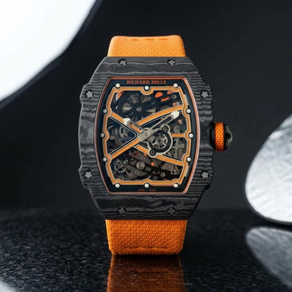 Black carbon tonneau watch with orange details on an orange textile strap, worn on the wrist below a navy suit cuff